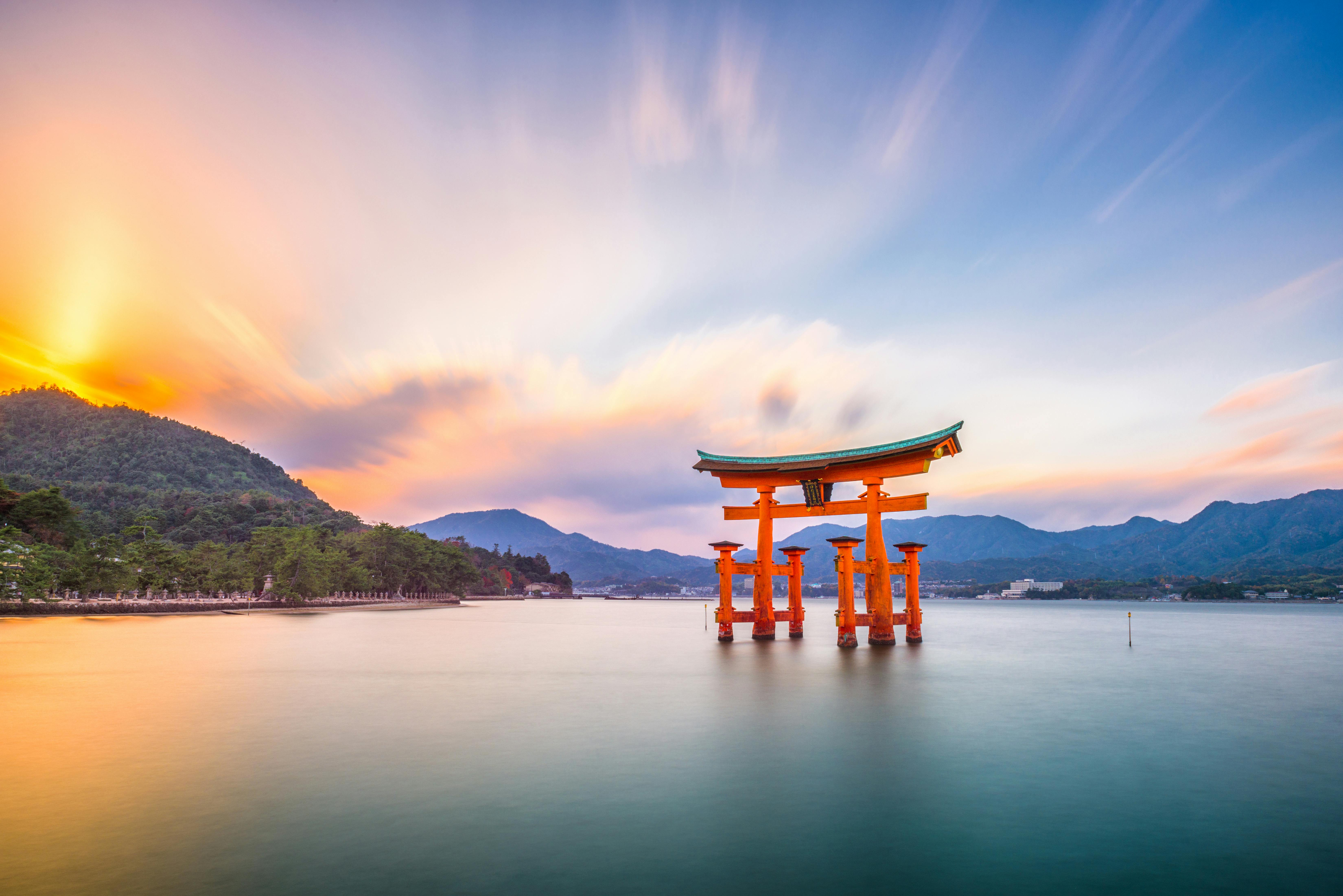 A large red torii gate stands in calm water near the shore, with mountains and a sunrise or sunset sky in the background at Itsukushima Shrine in Japan.