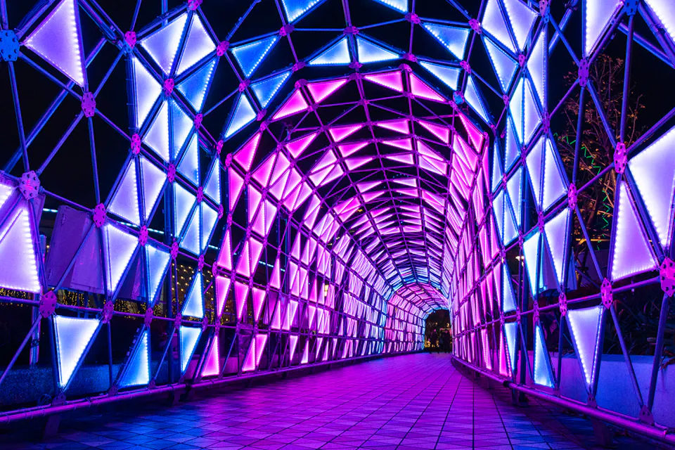 A tunnel with a geometric framework is illuminated by vibrant purple, pink, and blue lights, creating a visually striking pattern. The pathway beneath reflects the colorful glow, enhancing the tunnel's futuristic appearance.
