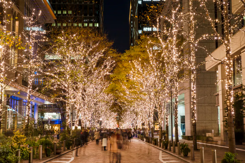 A bustling city street at night, lined with trees adorned in sparkling white lights. People walk along the illuminated path, surrounded by urban buildings. The scene is festive and vibrant.