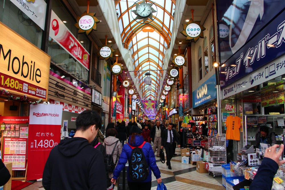 A busy indoor shopping street in Japan with many people walking, colorful shop signs, bright advertisements, and a high arched glass roof overhead.