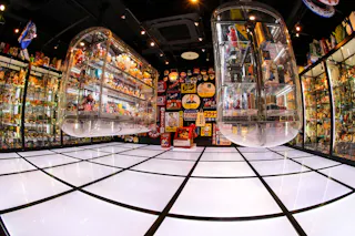 A brightly lit room with glass display cases filled with colorful toys and collectibles, and a glowing white tiled floor. The walls feature shelves and vibrant posters with Japanese text and cartoon characters.