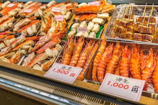 Fresh seafood displayed at a market, including prawns, crab legs, and skewered grilled seafood, with price tags in Japanese yen visible in front of each item.