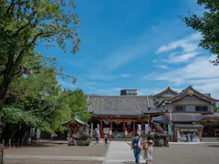 People walk toward a traditional Japanese temple with statues at the entrance, surrounded by trees and buildings, under a bright blue sky with scattered clouds.