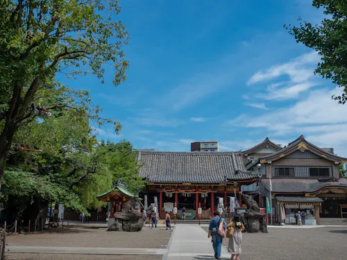 People walk toward a traditional Japanese temple with statues at the entrance, surrounded by trees and buildings, under a bright blue sky with scattered clouds.