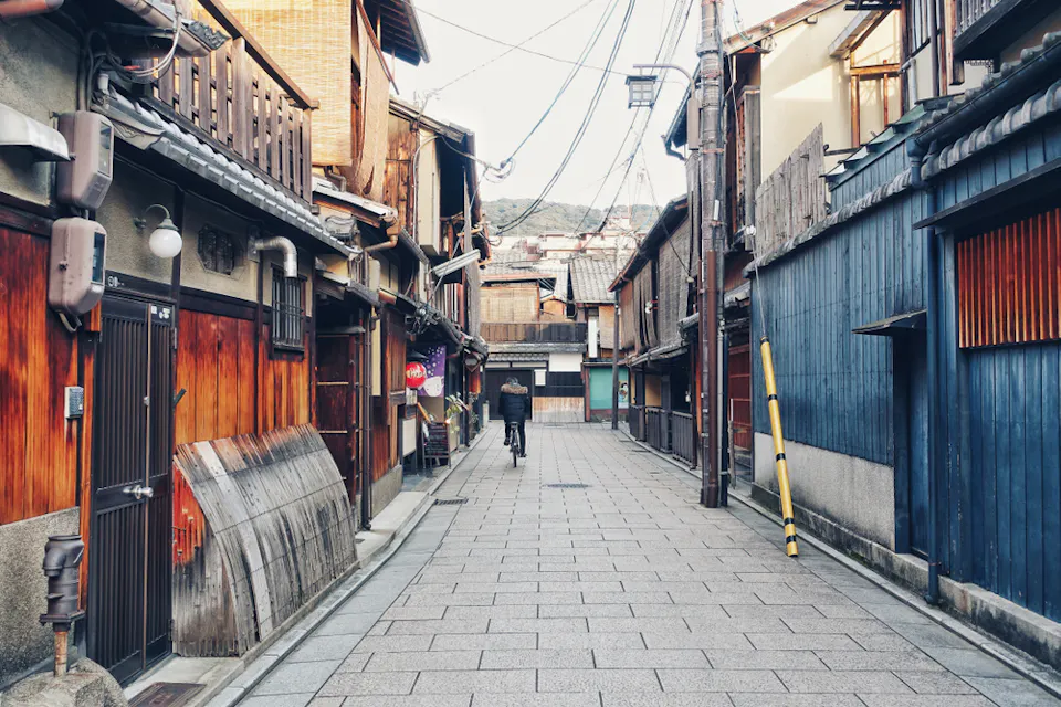 一条狭窄的传统日本街道上，有木制建筑、滑动门和瓦屋顶。一个人骑自行车走过，天空一片晴朗。头上的电线穿过街道，远处可见山脉。