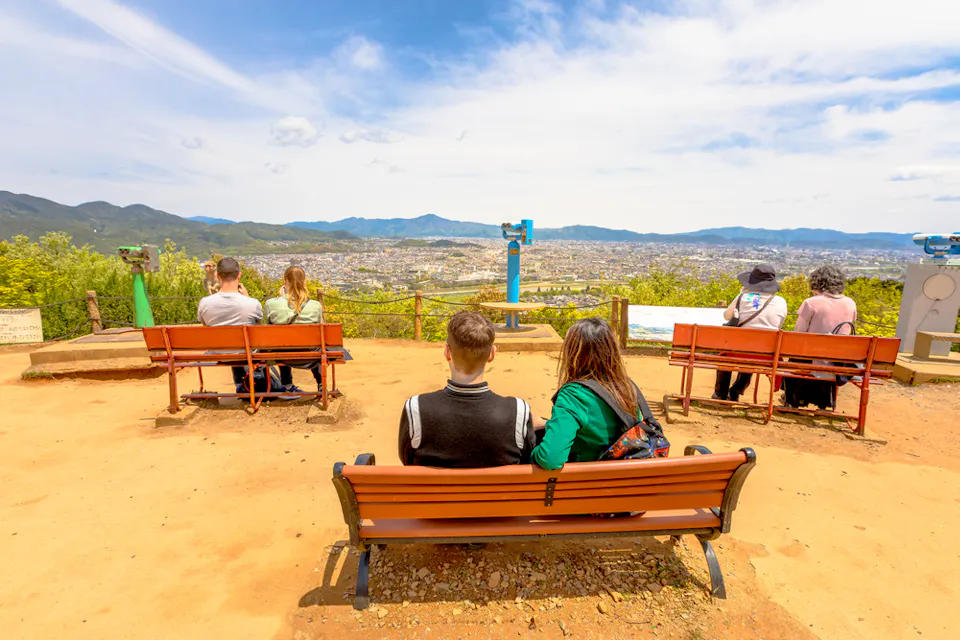 Popular Attraction Touristic Iwatayama Monkey Park Several people sit on wooden benches at a scenic overlook, with binocular stands in the foreground. They gaze out at a panoramic view of a sprawling city and mountainous landscape under a partly cloudy sky.