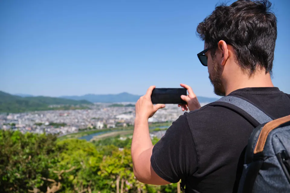 Tourist Taking a Photography of Kyoto City A person with dark hair and sunglasses is standing outdoors with a backpack on. They are using a smartphone to take a photo of the scenic view in front of them, which includes a city landscape and greenery under a clear blue sky.