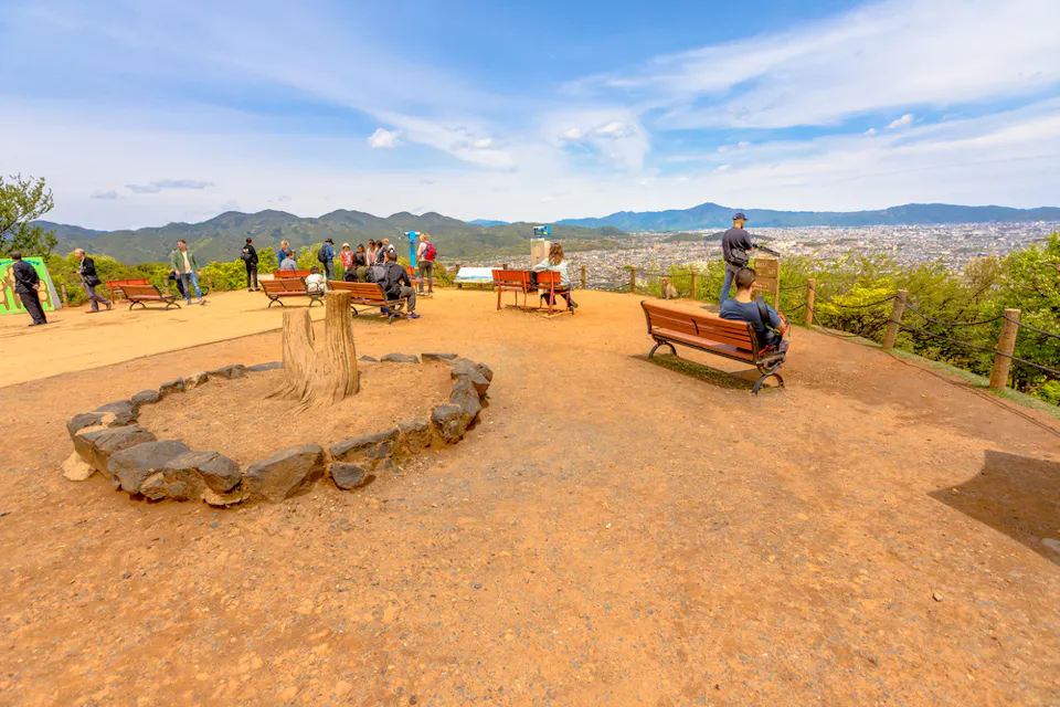 Touristic Attraction Iwatayama Monkey Park A scenic viewpoint on a hilltop with benches and a circular stone arrangement in the foreground. Visitors are sitting on benches and standing, enjoying the panoramic view of a city and mountains under a partly cloudy sky.
