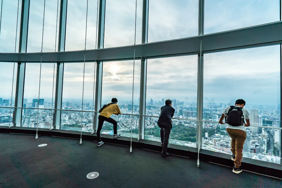 Three people are standing and leaning on the railing of a high observation deck with large windows, looking out at a vast urban landscape below. The sky is cloudy, and the cityscape stretches into the distance, covered in a light haze.