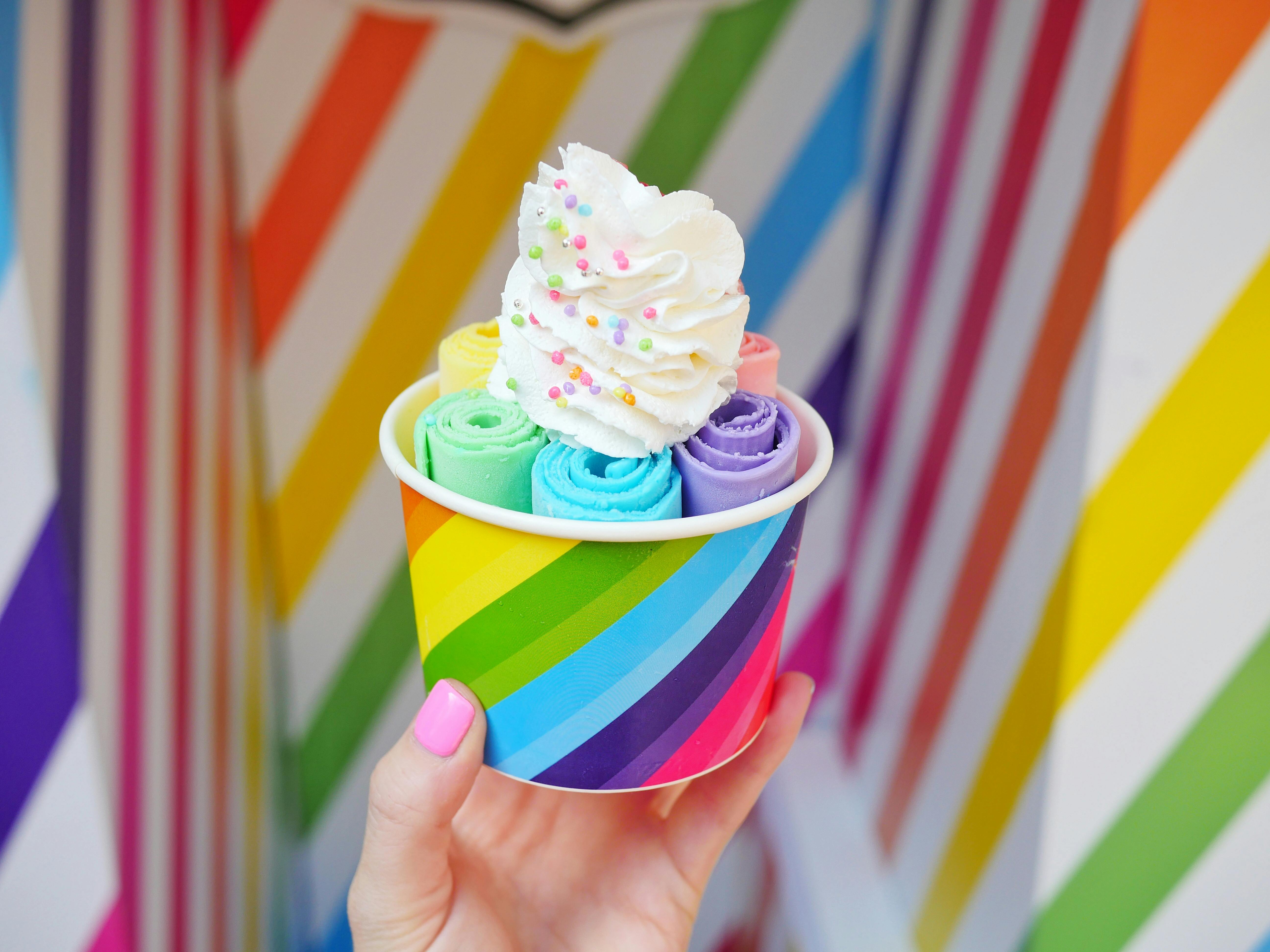 A hand holds a rainbow-striped cup filled with colorful rolled ice cream, topped with a swirl of whipped cream and rainbow sprinkles. The background features diagonal rainbow stripes.