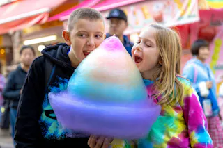 Two children excitedly share a giant, colorful cotton candy at an outdoor market, both about to take a bite. The background shows bright stalls and people, creating a lively atmosphere.