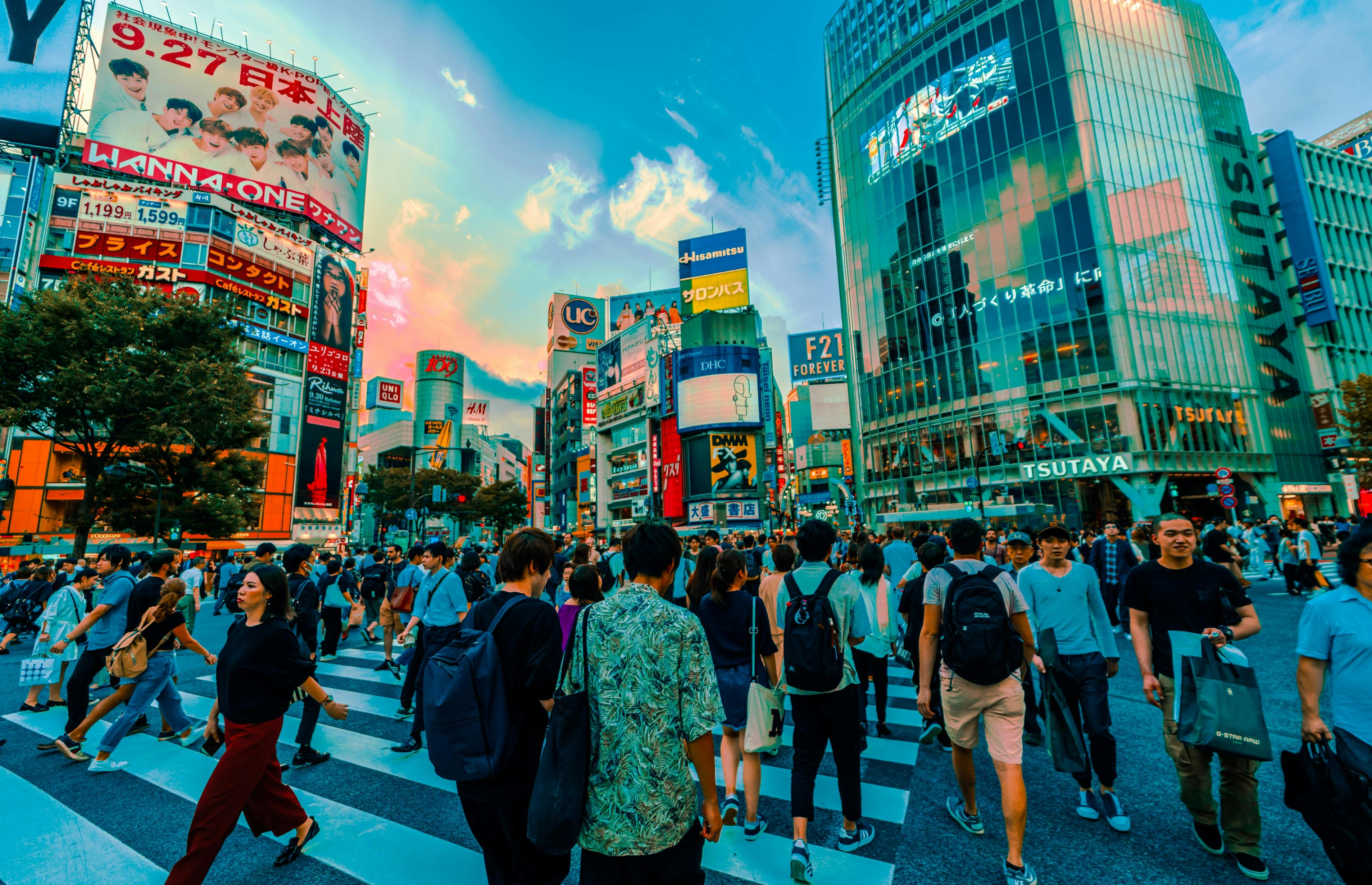 A busy crowd crosses the famous Shibuya Scramble intersection in Tokyo at sunset, surrounded by colorful billboards, tall buildings, and vibrant city lights.