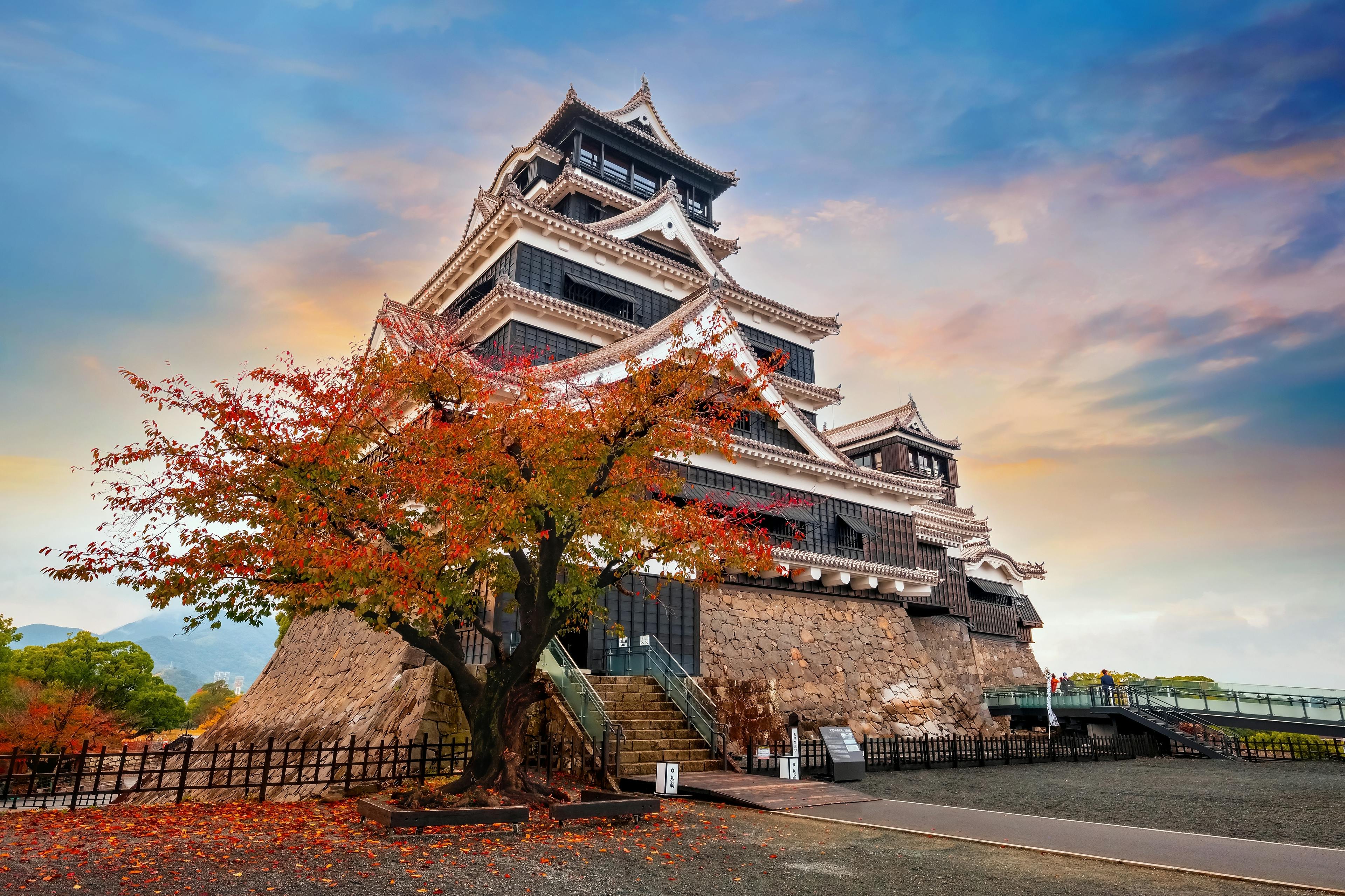 A traditional Japanese castle with tiered roofs stands behind a tree with red autumn leaves, set against a colorful sky at sunset. The castle is surrounded by stone walls and a fenced pathway.
