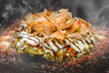 A close-up of a steaming okonomiyaki, a Japanese savory pancake, topped with drizzles of mayonnaise, okonomiyaki sauce, and bonito flakes on a hot griddle.