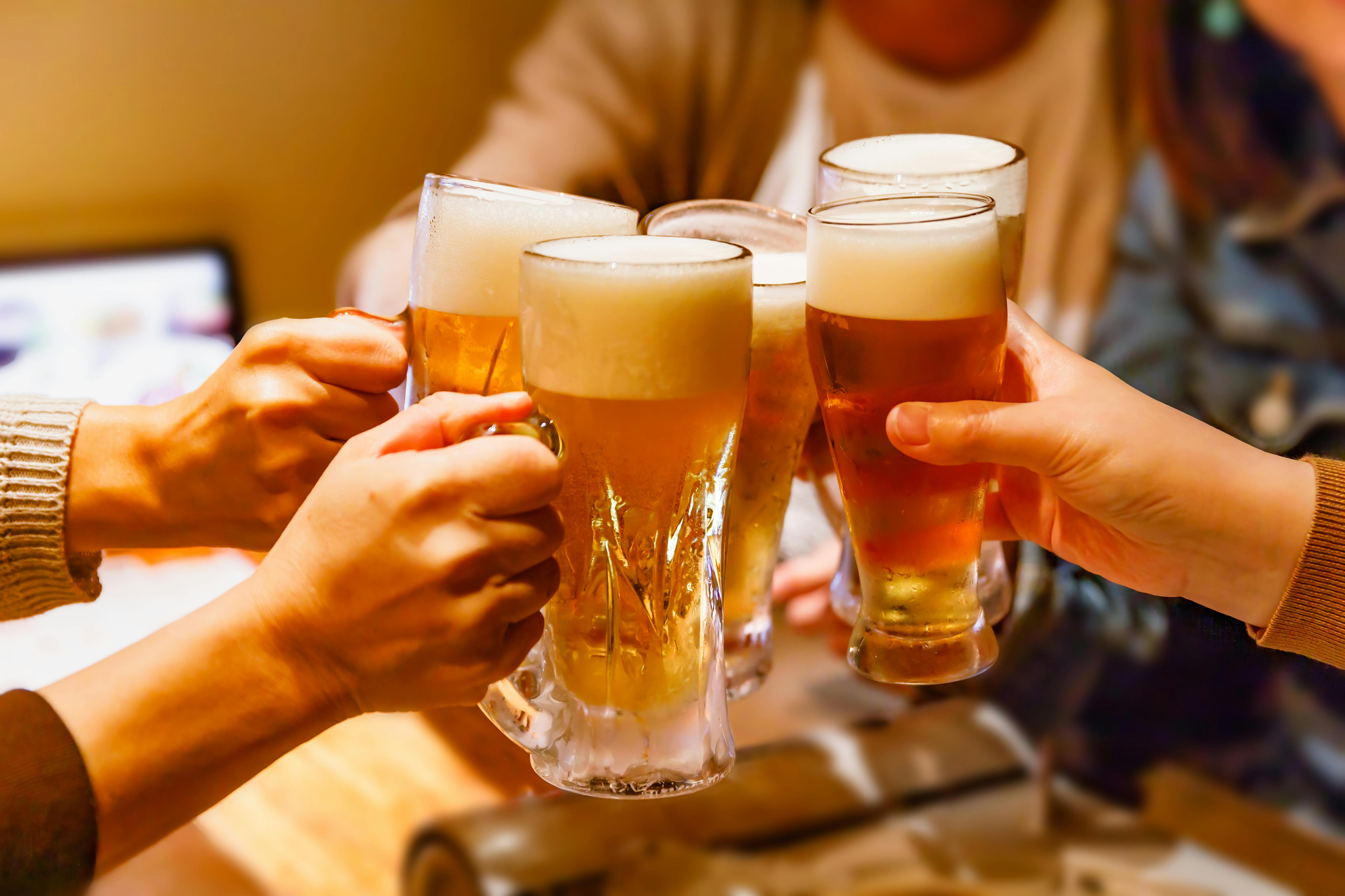 Four people clinking glasses of beer together in a celebratory toast, with foamy tops visible and a warm, indoor setting in the background.