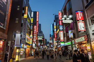 A busy city street in Japan at dusk, lined with colorful illuminated signs in Japanese, shops, and restaurants. Crowds of people walk along the street under the bright lights.