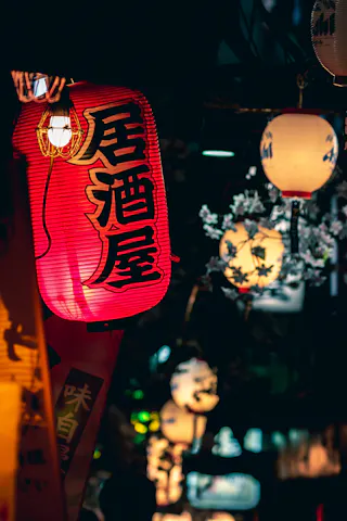 A vibrant red Japanese paper lantern with kanji characters hangs among other illuminated lanterns and cherry blossom decorations on a dimly lit street at night.