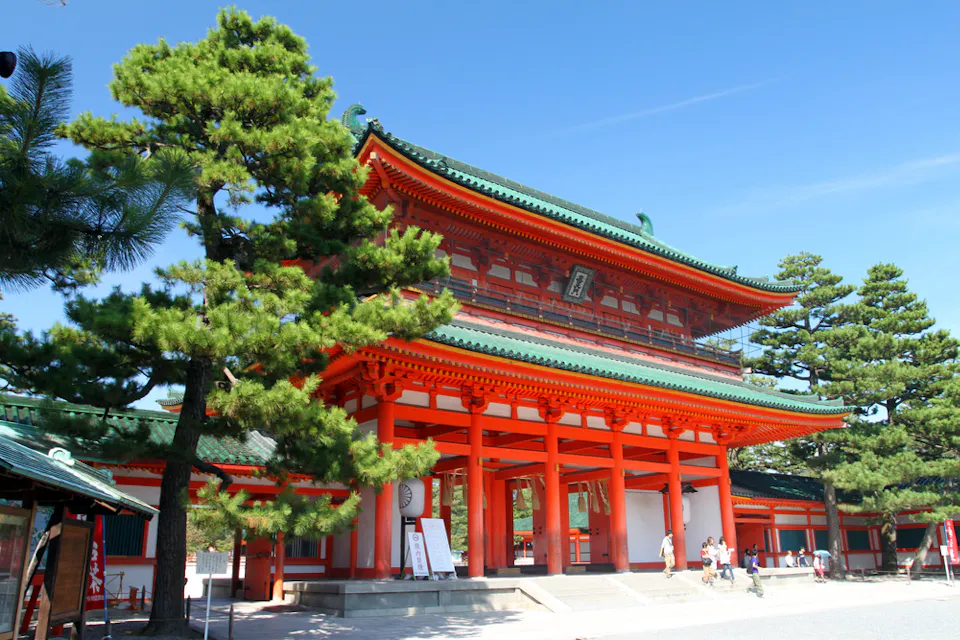 在清澈的蓝天下，传统的日本寺庙大门以鲜艳的橙色木材和绿色瓦顶显得格外突出，周围环绕着郁郁葱葱的绿色树木。一些人正在附近走动，为这座宏伟的建筑增添了尺寸感。