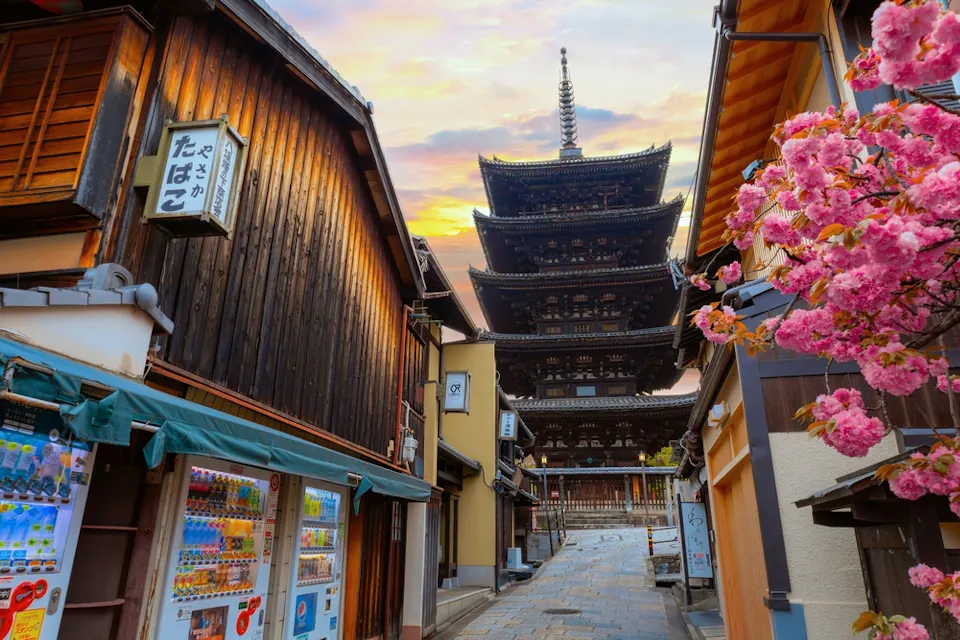 在京都的狭窄街道上，日落时分，古老的木制建筑和一台自动售货机映入眼帘。粉红色的樱花在图像右侧形成框架，通向背景中的标志性八坂塔，天空呈现出五彩斑斓的色彩。