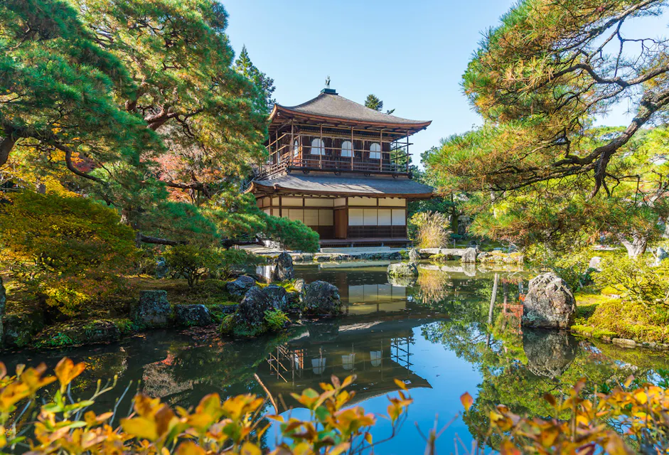 一个宁静的景象，京都的银阁寺（Ginkaku-ji）被郁郁葱葱的绿树环绕。亭子在平静的池塘中倒映，树木和岩石点缀着风景，营造出一个和平而和谐的场景。