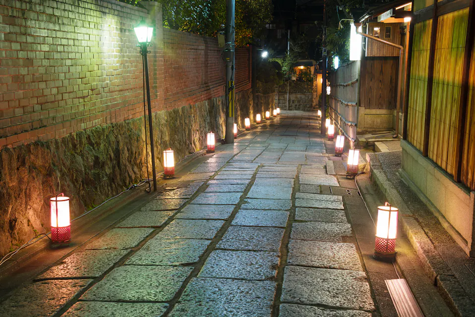 夜晚狭窄的石铺小巷，两侧点缀着传统的日本灯笼。小路左侧有砖墙，右侧是木结构，营造出宁静而诱人的氛围。