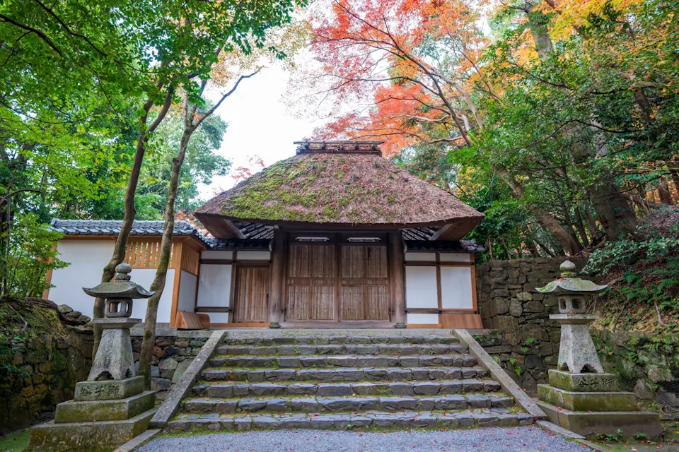 An ancient Japanese temple with moss-covered thatched roof situated amidst vibrant autumn foliage. Stone lanterns stand on either side of the mossy stone steps that lead up to the wooden entrance.