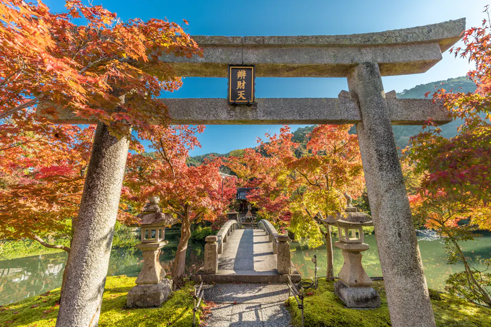 A stone torii gate surrounded by vibrant autumn foliage marks the entrance to a scenic Japanese garden. A path lined with stone lanterns leads across a small bridge over a tranquil pond, with a traditional structure visible in the background.