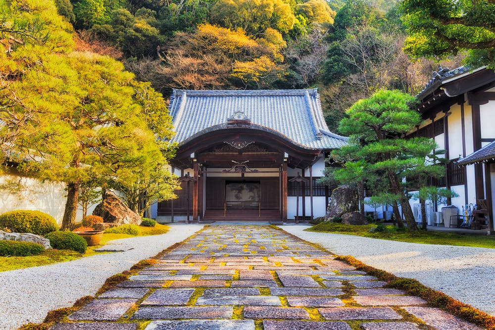 A serene Japanese temple with traditional wooden architecture, set amidst lush greenery. The temple is approached by a stone pathway lined with meticulously trimmed trees and shrubs. In the background, trees with autumn foliage enhance the tranquil scene.