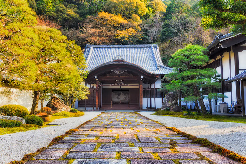 A serene Japanese temple with traditional wooden architecture, set amidst lush greenery. The temple is approached by a stone pathway lined with meticulously trimmed trees and shrubs. In the background, trees with autumn foliage enhance the tranquil scene.