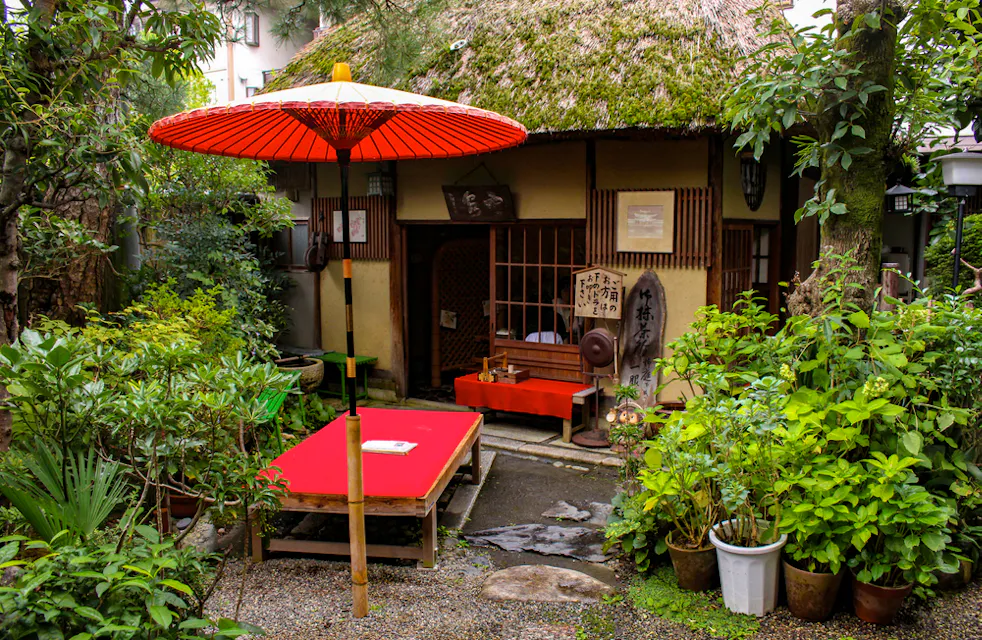 A quaint, traditional Japanese tea house surrounded by lush green plants. A red parasol stands over a red bench with a book on it. The thatched roof and wooden elements exude rustic charm, while stone paths and potted plants create a serene atmosphere.