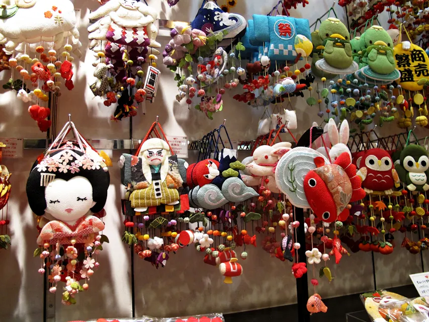 A display of various colorful Japanese ornaments made of fabric. The decorations include figures like a geisha, an old man, animals, fish, and flowers. Each ornament is adorned with intricate details, hanging just above eye level at a market stall.