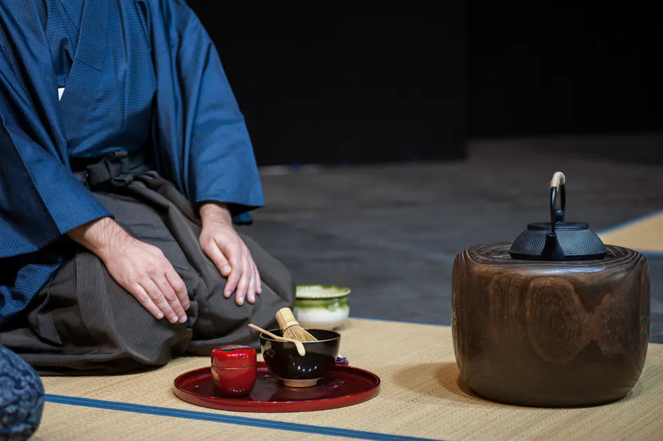 A person dressed in traditional Japanese attire sits on a tatami mat with hands resting on their knees. In front of them are various tea ceremony items, including a black tea kettle on a wooden stand, a red bowl, and a whisk, all arranged on a tray.