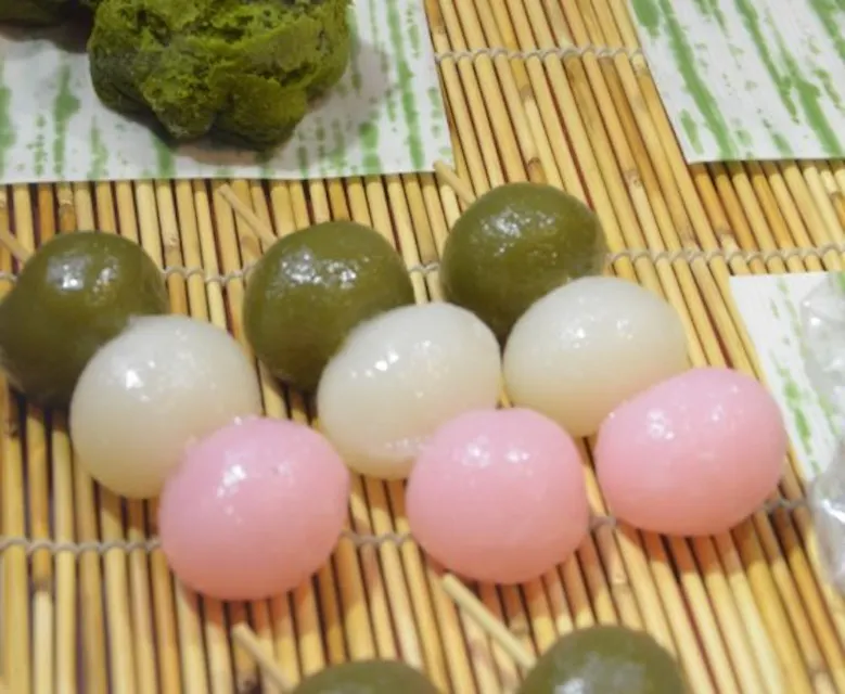 Dango A close-up of colorful dango skewers resting on a bamboo mat. The skewers feature green, white, and pink rice dumplings. In the background, there is a small pile of green tea-flavored sweets.