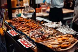 A variety of skewered meats and seafood are displayed on a market food stall, with grilled items arranged neatly on trays and in foil, ready to be served. Signs with prices are visible next to the food.