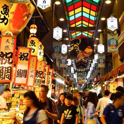 Kyoto Nishiki Market Food Tour A bustling indoor market in Japan with brightly colored signs in Japanese hanging from the ceiling. Numerous people walk through the market, browsing various food stalls. The ceiling has a stained-glass design, creating a vibrant and lively atmosphere.