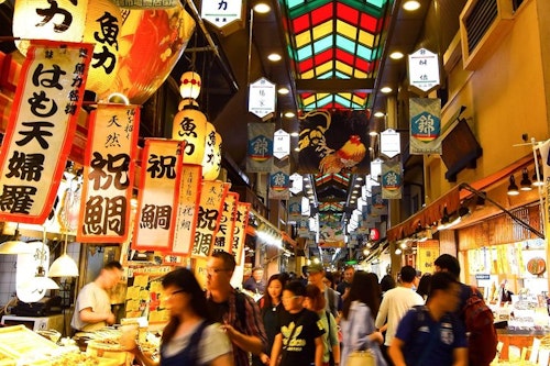 A bustling indoor market in Japan with brightly colored signs in Japanese hanging from the ceiling. Numerous people walk through the market, browsing various food stalls. The ceiling has a stained-glass design, creating a vibrant and lively atmosphere.