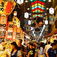 Kyoto Nishiki Market Food Tour A bustling indoor market in Japan with brightly colored signs in Japanese hanging from the ceiling. Numerous people walk through the market, browsing various food stalls. The ceiling has a stained-glass design, creating a vibrant and lively atmosphere.