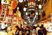 A bustling indoor market in Japan with brightly colored signs in Japanese hanging from the ceiling. Numerous people walk through the market, browsing various food stalls. The ceiling has a stained-glass design, creating a vibrant and lively atmosphere.
