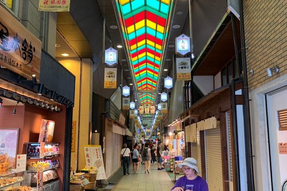 Kyoto Nishiki Market Food Tour A vibrant indoor market with a colorful stained-glass ceiling. Stalls line both sides, showcasing various goods. People are walking through, and a person in a purple shirt and hat is in the foreground. Signs with Japanese text hang from above.