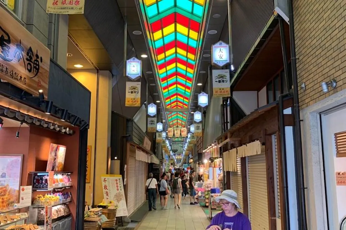 A vibrant indoor market with a colorful stained-glass ceiling. Stalls line both sides, showcasing various goods. People are walking through, and a person in a purple shirt and hat is in the foreground. Signs with Japanese text hang from above.