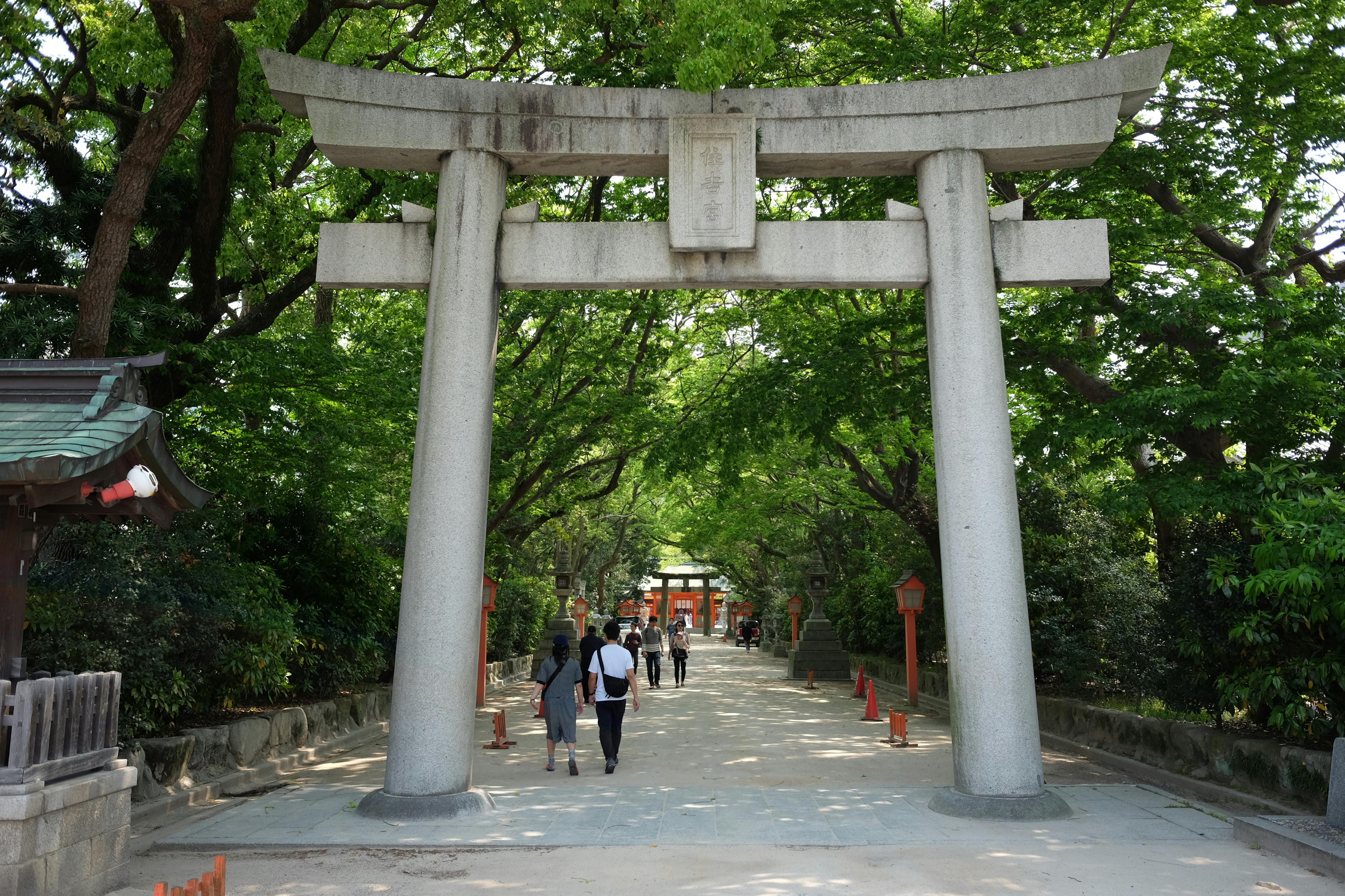 A large stone torii gate stands at the entrance to a tree-lined path, with several people walking toward a distant shrine under a canopy of green leaves.
