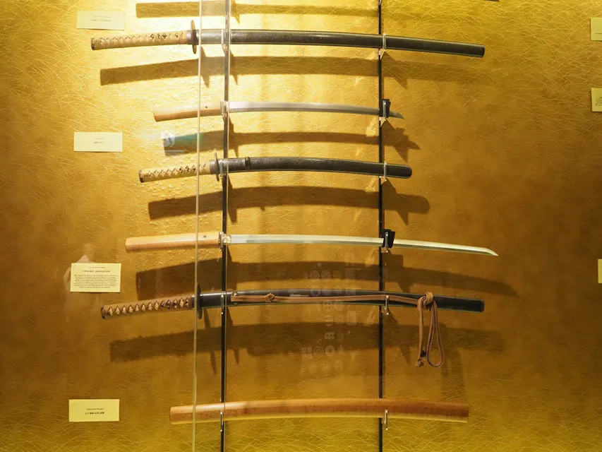 텍스처가 있는 노란 벽에 수직으로 장착된 여섯 개의 일본 검이 전시되어 있습니다. 각 검은 독특한 디자인을 가지며, 손잡이와 칼집 스타일이 다양합니다. 각 검 옆에 정보가 적힌 설명 레이블이 배치되어 있습니다. 전시는 잘 조명되어 세부 사항을 강조합니다.