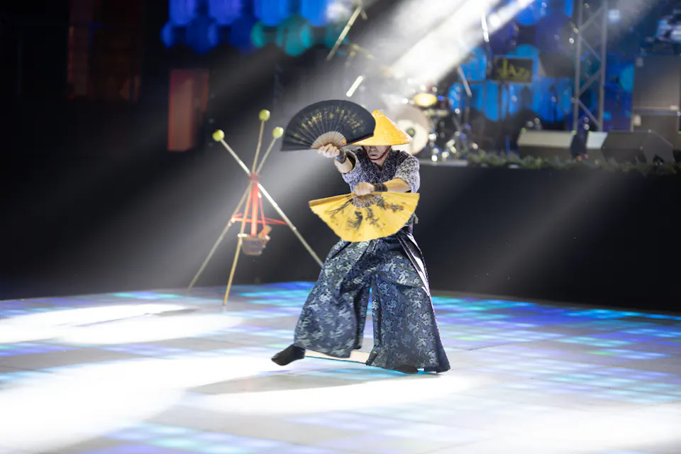 전통 의상을 입고 투구 모자와 흐르는 바지를 착용한 댄서가 밝게 조명된 무대에서 공연을 펼치고 있다. 그들은 두 개의 큰 검은색과 노란색 부채를 들고 능숙하게 조작하고 있다. 배경에는 악기와 무대 조명이 보인다.