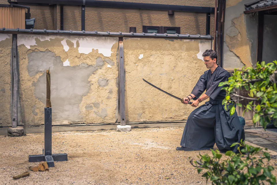 전통 일본 복장을 한 남성이 카타나로 무술을 연습하고 있다. 그는 나무로 된 표적을 향해 서 있는 모래 마당에 있다. 배경에는 낡고 질감 있는 벽과 건물의 일부분이 보인다. 전경에는 녹색 나뭇잎이 있다.