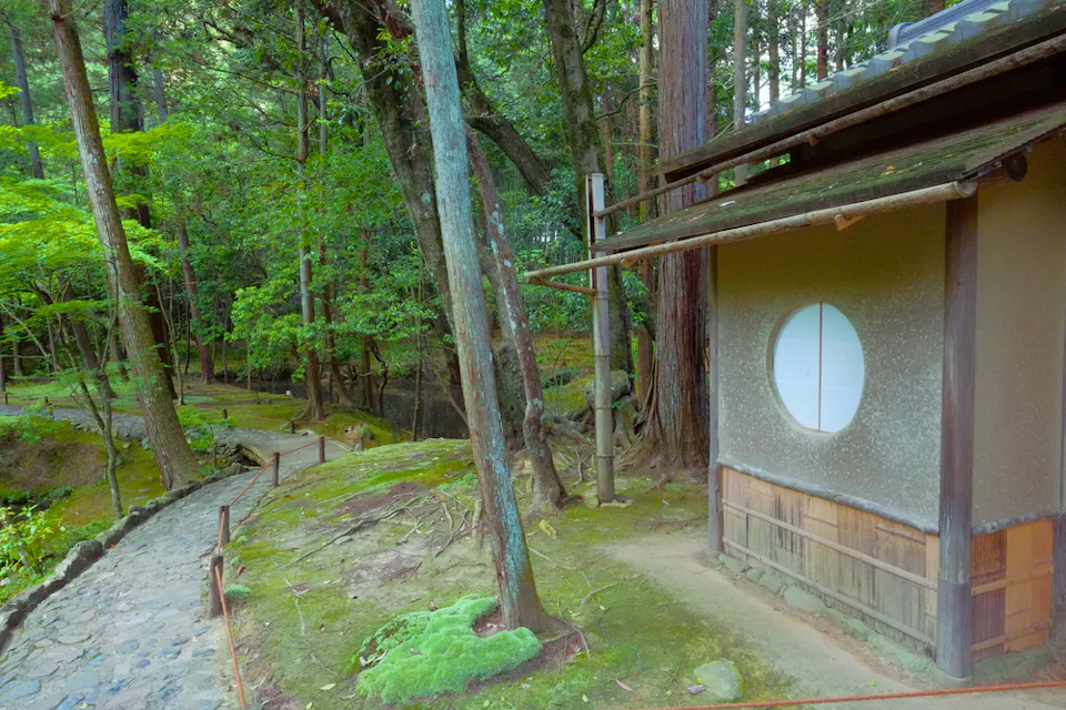 高い木々と緑豊かな環境に囲まれた、丸い窓を持つ伝統的な日本の茶室。木製の柱に沿った石の小道が家の横を通り抜ける。苔むした地面と樹の根が、静かで自然な雰囲気を引き立てている。