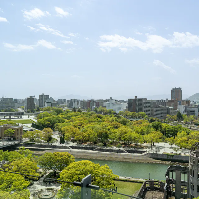 Orizuru Tower View A cityscape view of Hiroshima, Japan, with green trees, a river, modern buildings, and the Hiroshima Peace Memorial (Atomic Bomb Dome) in the foreground under a partly cloudy sky.