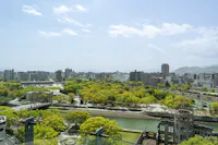 A cityscape view of Hiroshima, Japan, with green trees, a river, modern buildings, and the Hiroshima Peace Memorial (Atomic Bomb Dome) in the foreground under a partly cloudy sky.