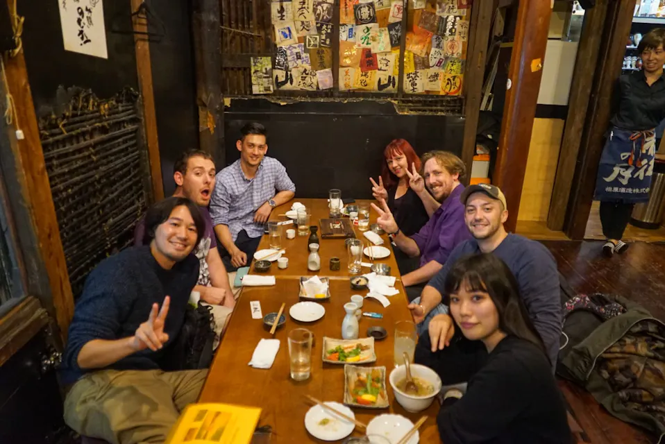 A group of seven people sitting at a wooden table in a cozy restaurant, sharing a meal. The table is filled with various dishes and drinks. Some individuals are smiling and making peace signs. The walls are decorated with posters and memorabilia, creating a vibrant atmosphere.