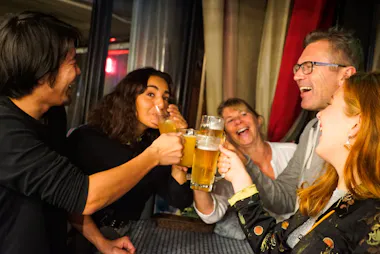 Five people are smiling and laughing while clinking glasses of beer together at a table, appearing to enjoy a lively social gathering indoors.