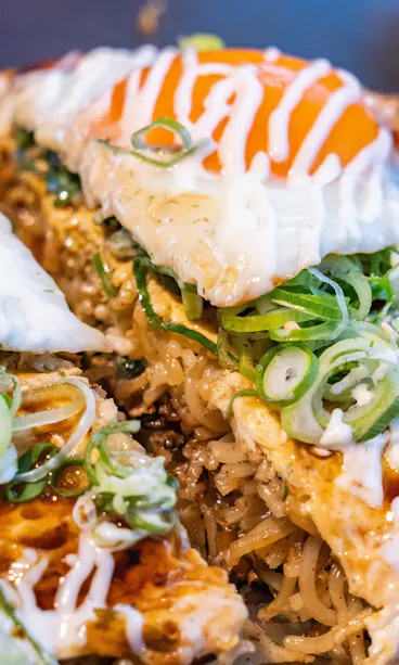 Okonomiyaki Close-up of Japanese okonomiyaki topped with fried eggs, sliced green onions, mayonnaise, and savory sauce, showing vibrant colors and textures of the ingredients.